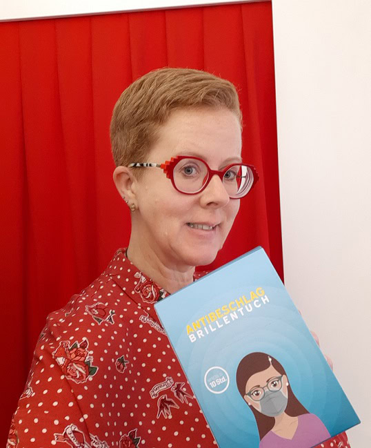 Person hält Antibeschlag-Brillentuch vor rotem Hintergrund.